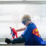 A young boy dressed as a superhero, playing with a toy by a window, embracing his imagination.